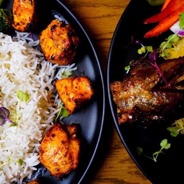 Two black plates of Indian food, one with chicken tikka and rice, the other with lamb chops.