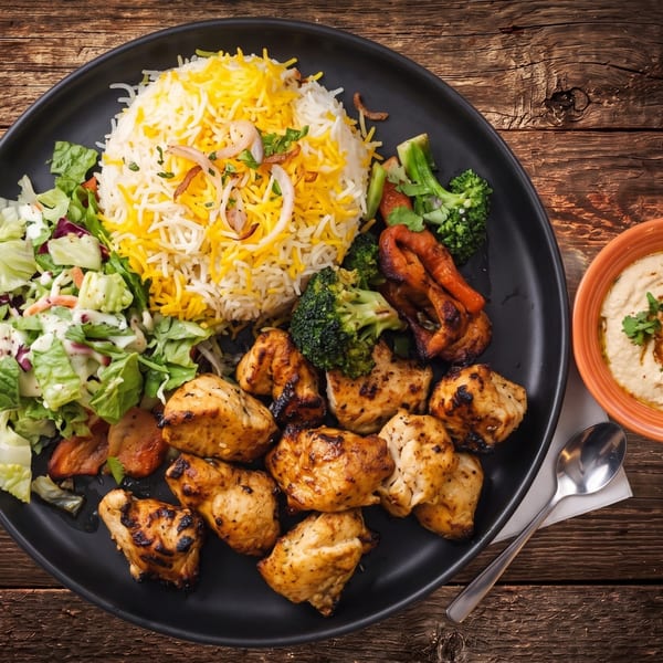 Grilled chicken pieces, yellow rice, salad, and roasted vegetables on a black plate with hummus.