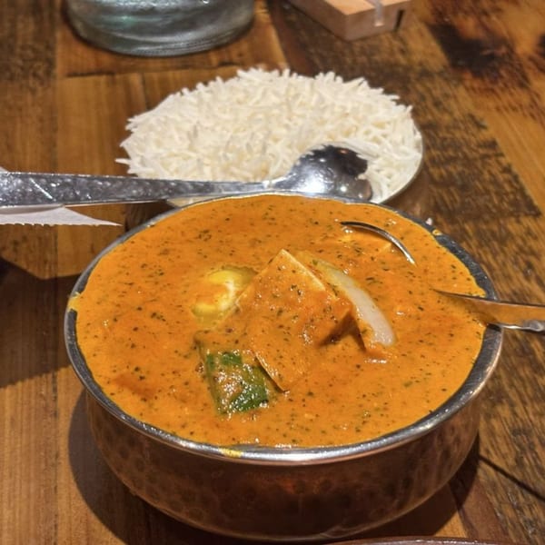 A bowl of orange curry with paneer and vegetables, served with a side of white rice.