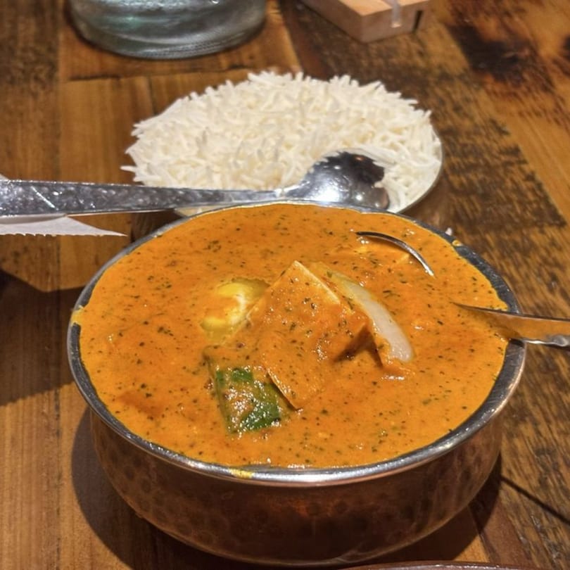 A bowl of orange curry with paneer and vegetables, served with a side of white rice.