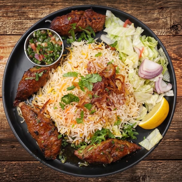 A black plate of biryani rice, grilled chicken, fresh salad, and a lemon wedge on a wooden table.